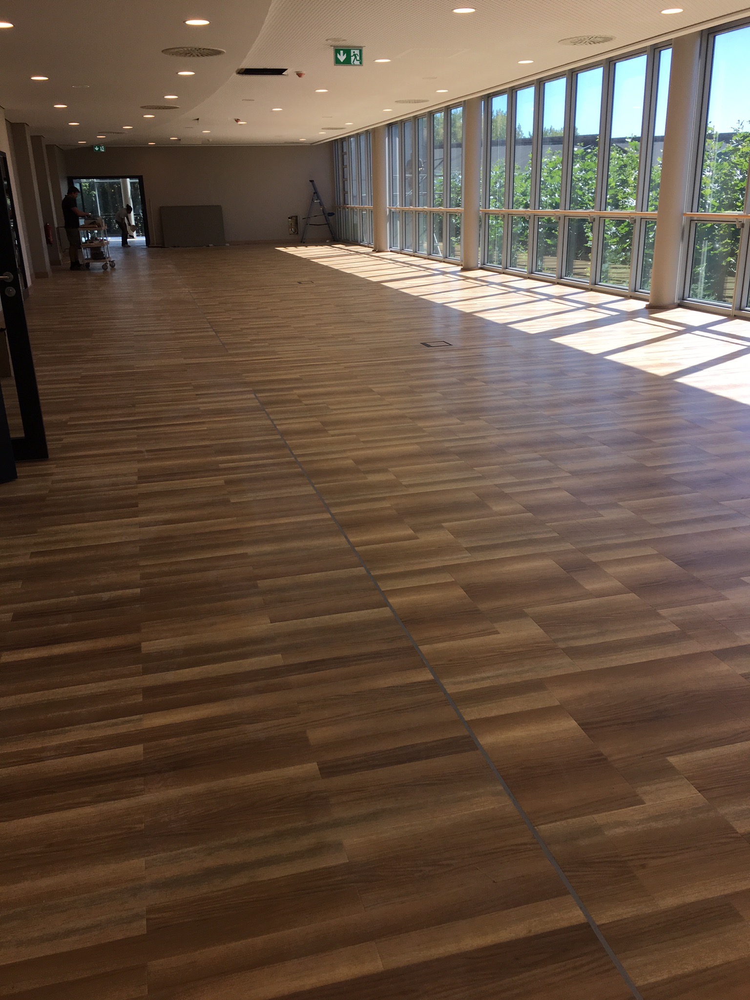 Nowa, brązowa podłoga z paneli ułożona w przestronnym, jasnym pomieszczeniu z dużymi oknami i słupami, widoczny wózek narzędziowy i drabina w tle.