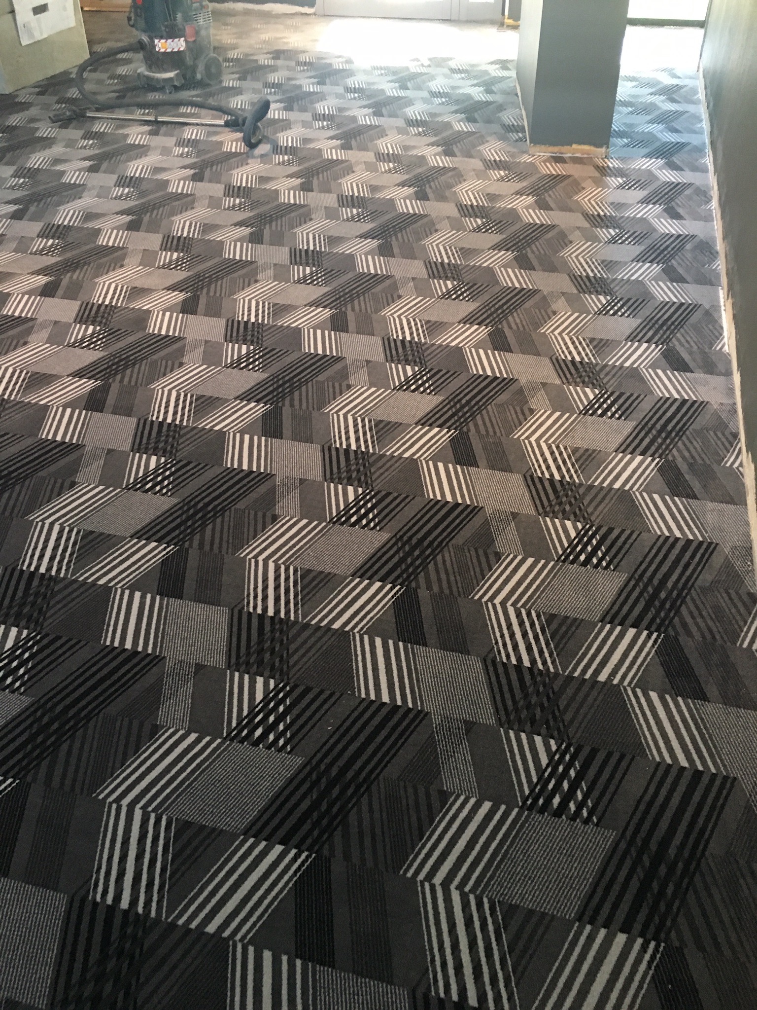 Układanie wykładziny dywanowej w geometryczny wzór w odcieniach szarości, widoczny odkurzacz przemysłowy w tle.