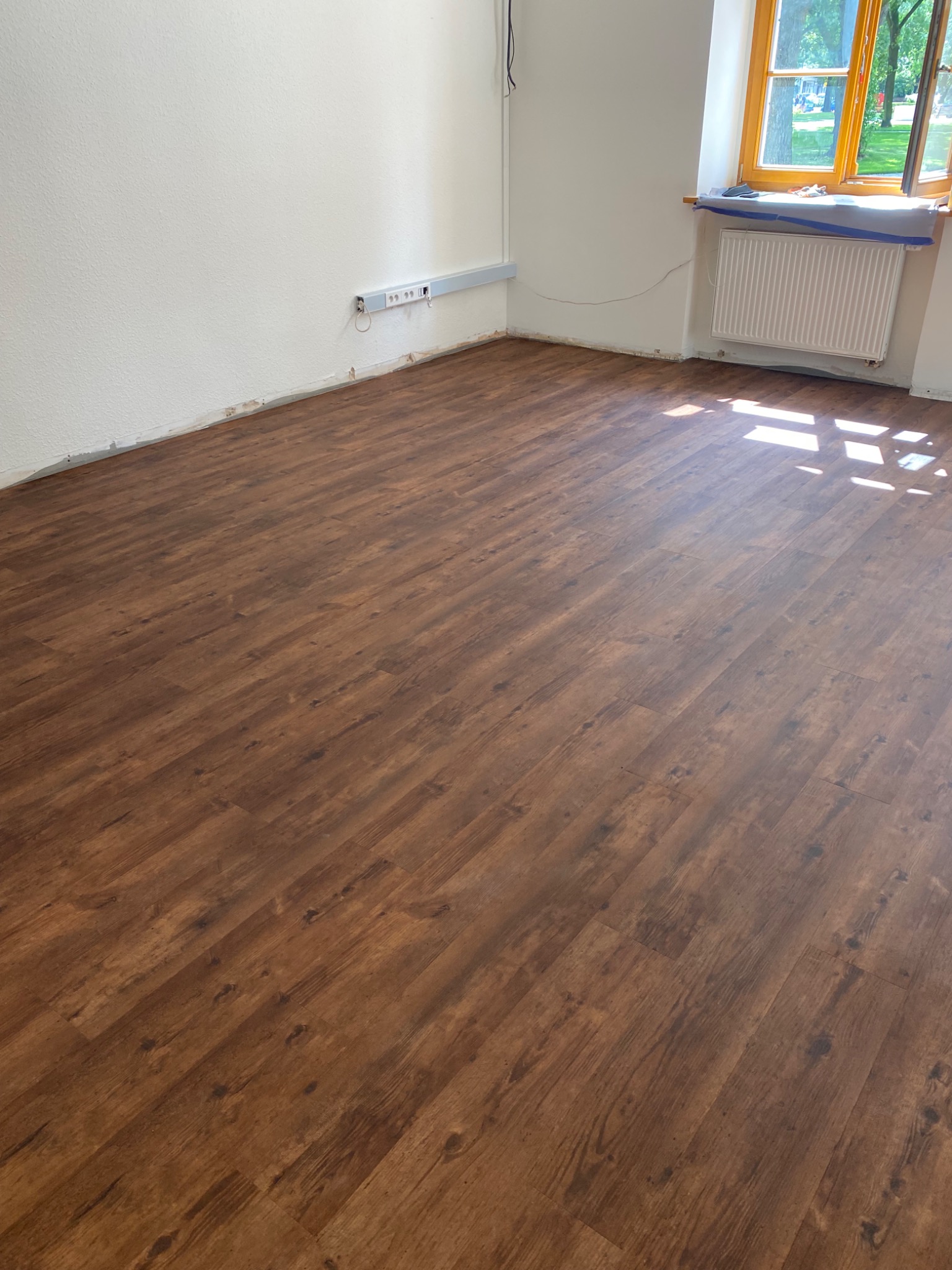 Nowo położona podłoga z paneli w odcieniach brązu z widocznym wzorem drewna, jasne ściany i okno z widokiem na zieleń na zewnątrz, listwy przypodłogowe jeszcze nie zamontowane.