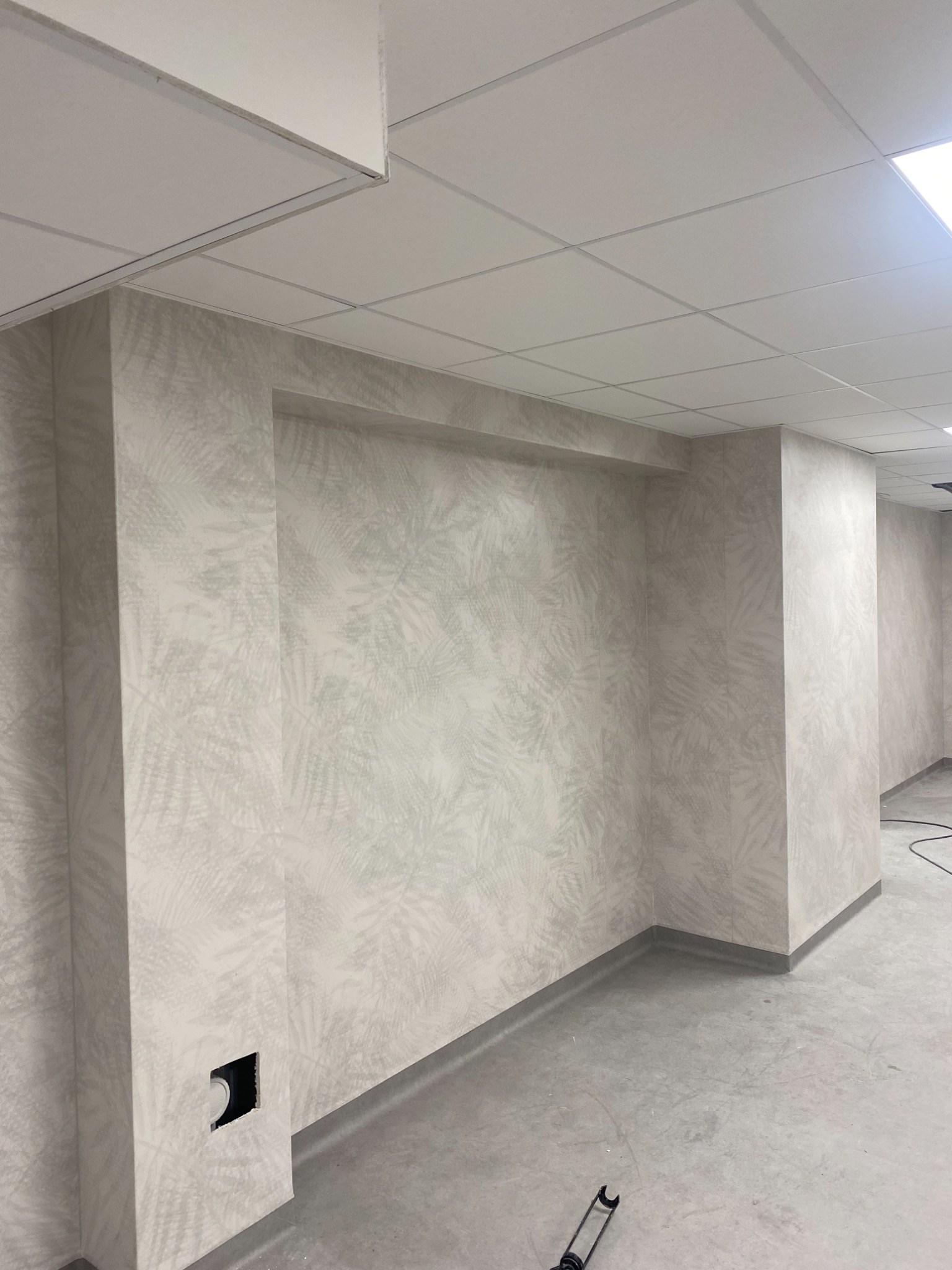 Wykończone wnętrze z wykładziną ścienną w delikatny wzór liści, widoczne panele sufitowe i szara listwa przypodłogowa, surowa podłoga, w tle kabel i narzędzie.