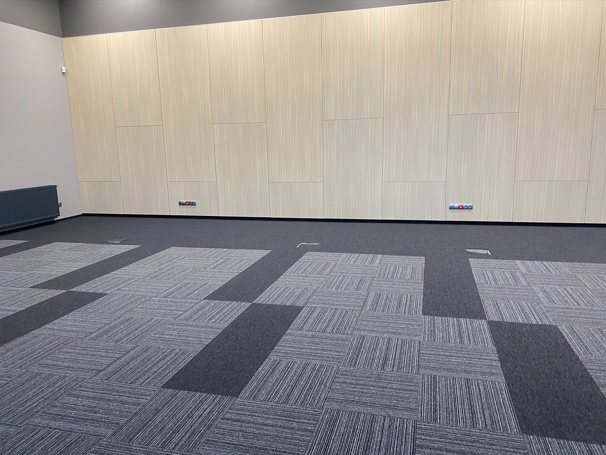 Wnętrze biurowe z szarą wykładziną dywanową w geometryczny wzór, jasne panele ścienne, grzejnik i gniazdka elektryczne przy podłodze.