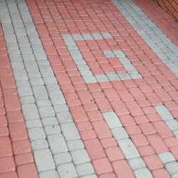 Ułożona kostka brukowa w odcieniach czerwieni i szarości, tworząca wzór. Widoczny fragment ściany z cegły i odpływ liniowy. Perspektywa z góry.