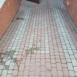 Umyta część kostki brukowej przed garażem, widoczne różnice w kolorze, ceglana ściana po lewej stronie. Częściowo otwarte, brązowe drzwi garażowe.