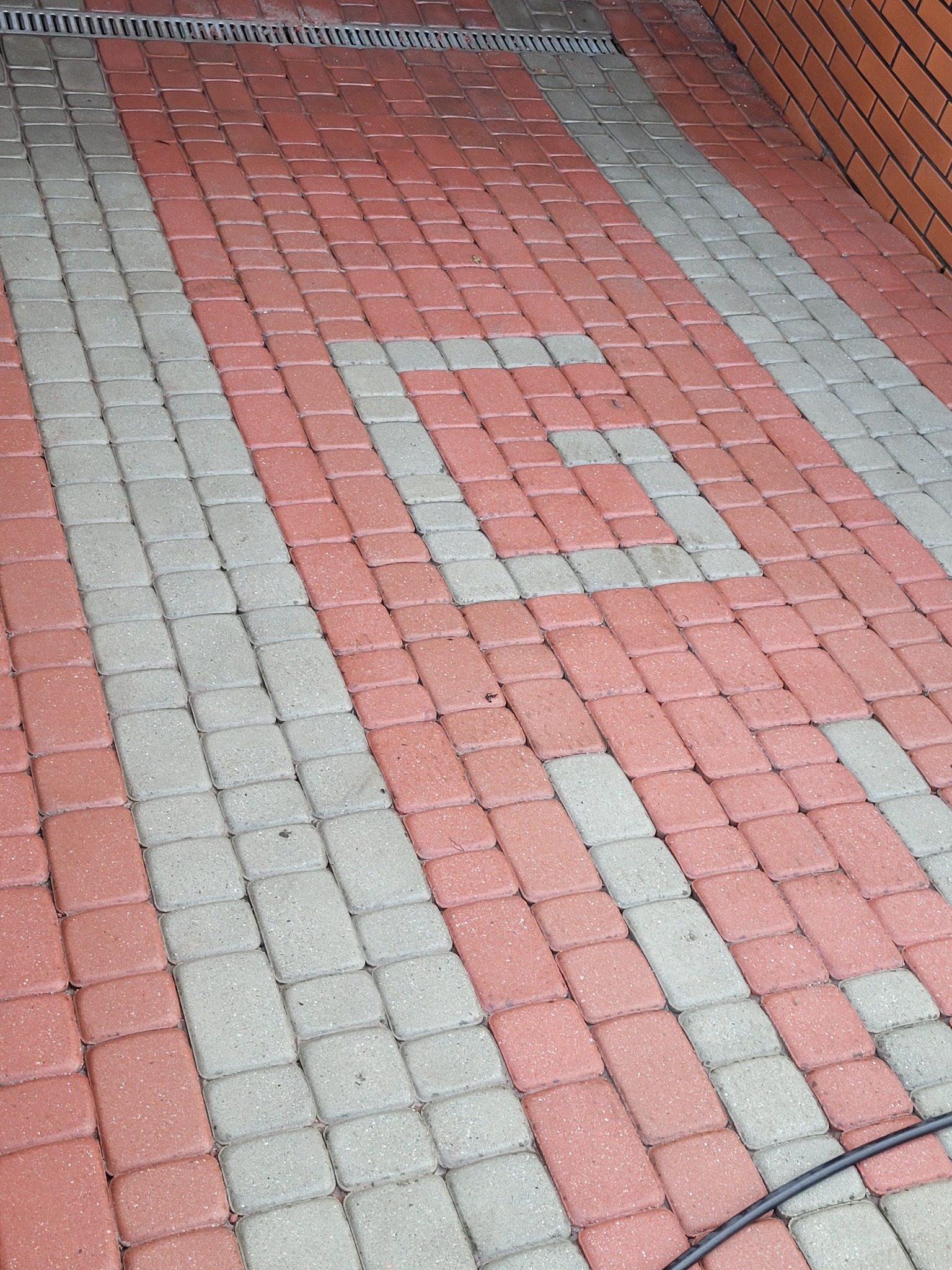 Ułożona kostka brukowa w odcieniach czerwieni i szarości, tworząca wzór. Widoczny fragment ściany z cegły i odpływ liniowy. Perspektywa z góry.