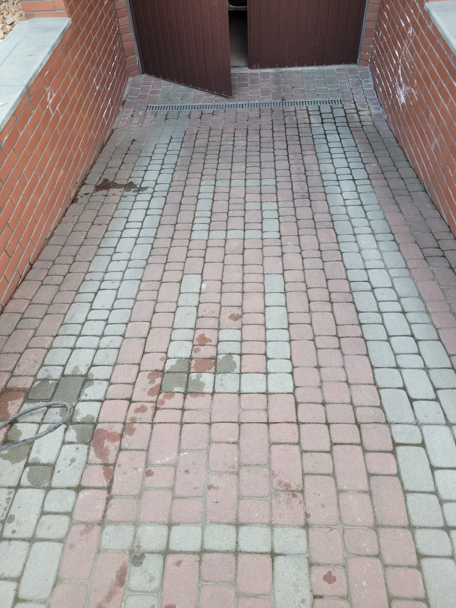 Umyta część kostki brukowej przed garażem, widoczne różnice w kolorze, ceglana ściana po lewej stronie. Częściowo otwarte, brązowe drzwi garażowe.