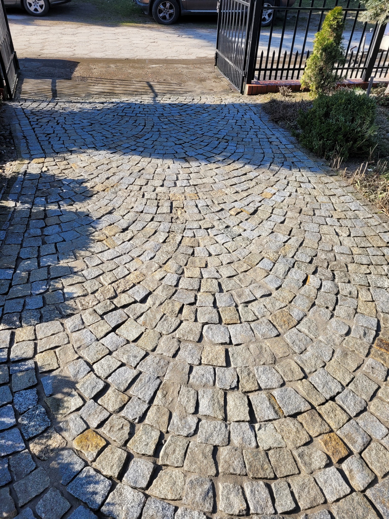 Ułożona w łuk kostka brukowa w odcieniach szarości i beżu, prowadząca do bramy wjazdowej. Widoczne cienie drzew i krzewów. Słoneczny dzień.