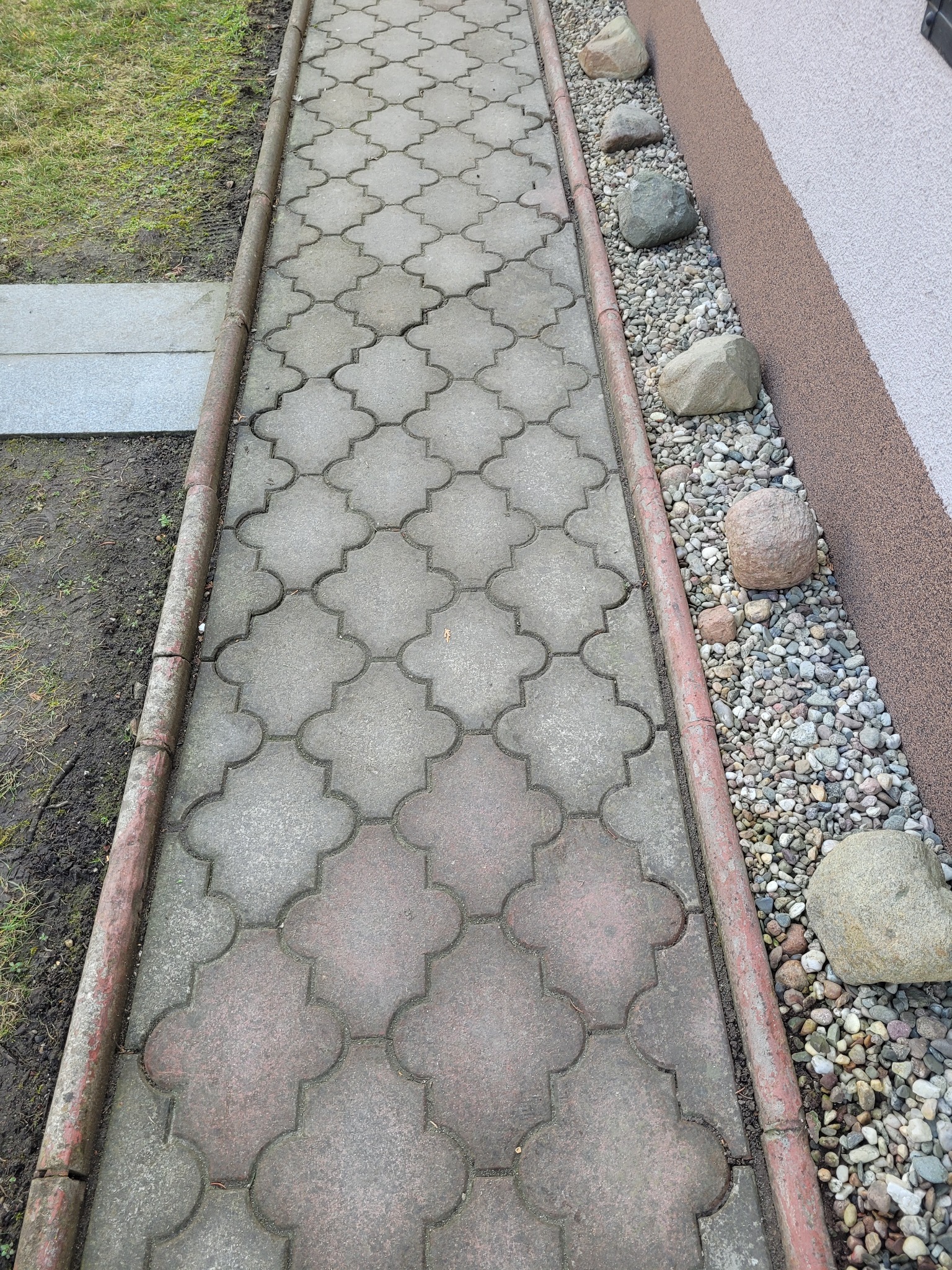 Ułożona kostka brukowa w geometryczny wzór, obrzeża z czerwonej cegły, kamienie dekoracyjne przy elewacji budynku. Widok z góry na ścieżkę.