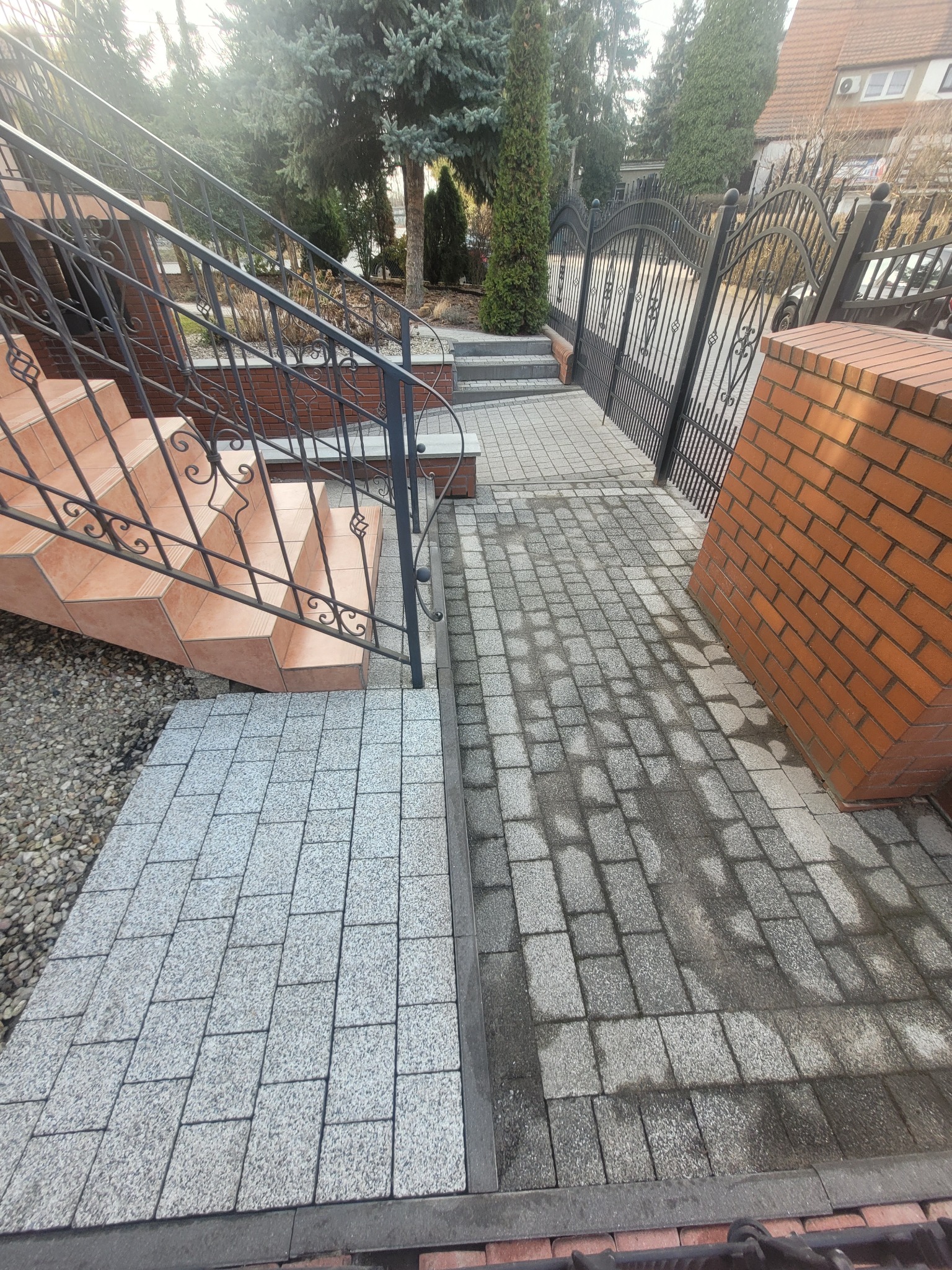 Częściowo umyta kostka brukowa na podjeździe, kontrastująca z brudną powierzchnią. Schody z terakoty i metalowa balustrada. Widoczne ogrodzenie i zieleń w tle.