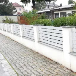 Nowoczesne białe ogrodzenie z poziomych paneli i szarymi elementami betonowymi, widok z chodnika z kostki brukowej w pochmurny dzień.