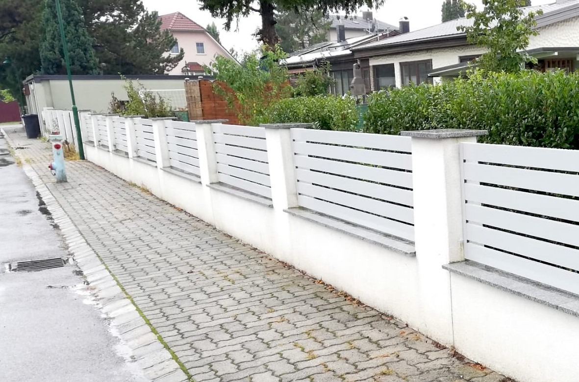 Nowoczesne białe ogrodzenie z poziomych paneli i szarymi elementami betonowymi, widok z chodnika z kostki brukowej w pochmurny dzień.