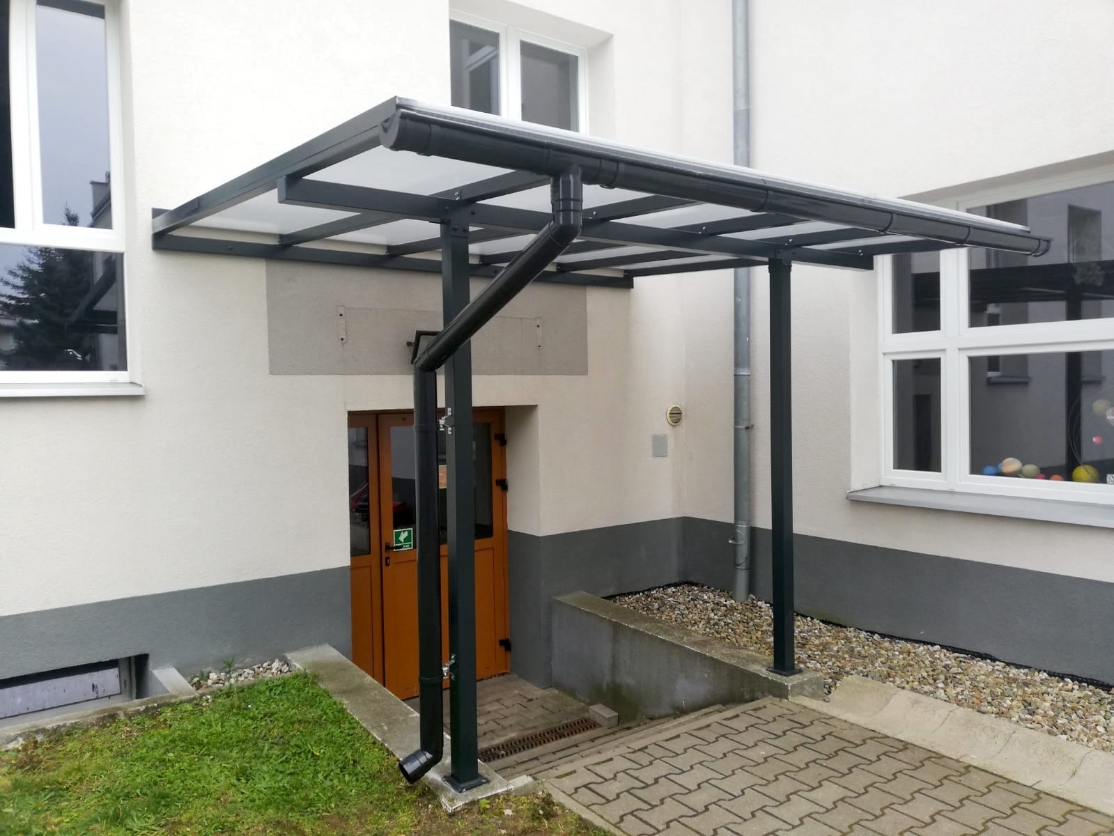 Zadaszenie nad wejściem do budynku, konstrukcja metalowa z rynnami, wsparta na słupach, widok z zewnątrz, kostka brukowa na podłożu.