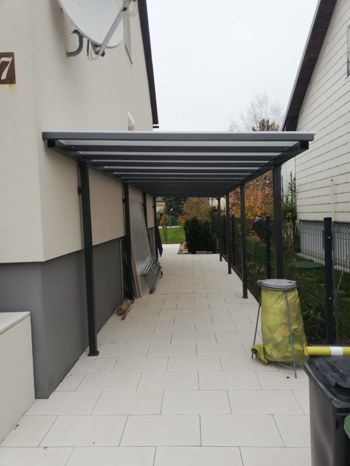 Metalowa wiata przydomowa z płaskim dachem, wsparta na słupach, osłaniająca przejście wyłożone jasną kostką brukową, widok z perspektywy przechodnia.