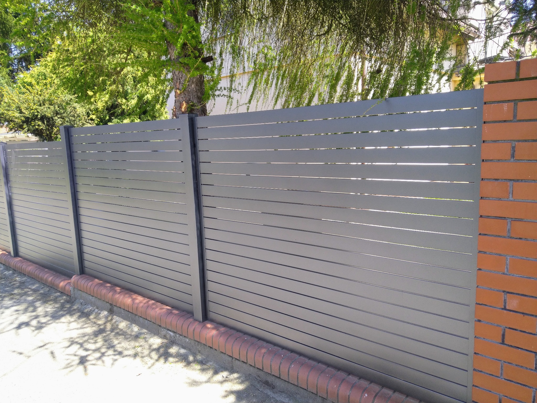 Nowoczesne ogrodzenie panelowe z poziomych listew w kolorze szarym, z słupkami i podmurówką z cegły klinkierowej, na tle zieleni drzew.