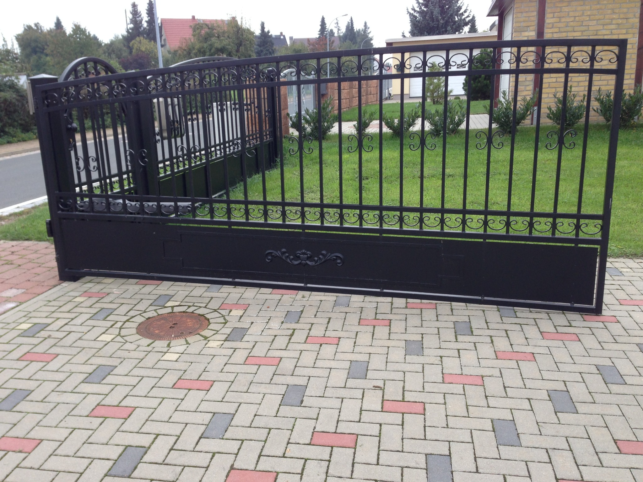 Czarna, przesuwna brama wjazdowa z kutymi elementami dekoracyjnymi, ustawiona na kostce brukowej w kolorach szarości, czerwieni i bieli, z widoczną studzienką kanalizacyjną w pobliżu.