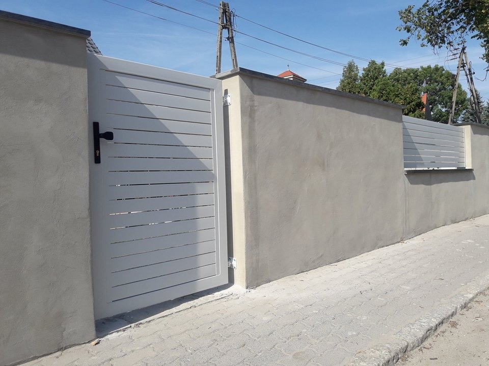 Nowoczesna brama wjazdowa z poziomych, jasnych paneli, wkomponowana w szary, otynkowany mur ogrodzeniowy z analogicznymi panelami w górnej części. Czarny, prosty uchwyt na bramie. Widoczne słupy...