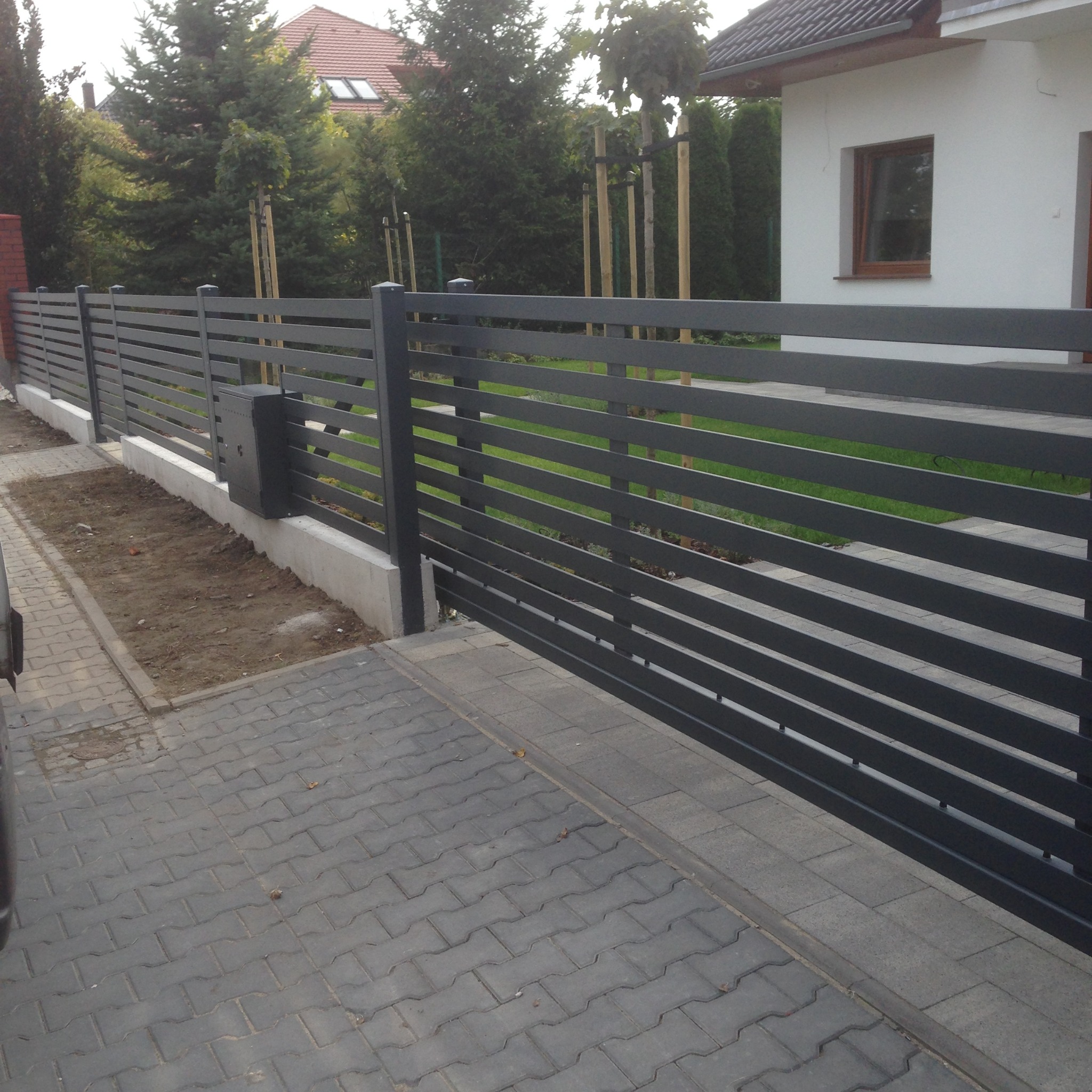 Nowoczesne, poziome ogrodzenie metalowe w kolorze grafitowym, zautomatyzowana brama wjazdowa, kostka brukowa przed domem jednorodzinnym.
