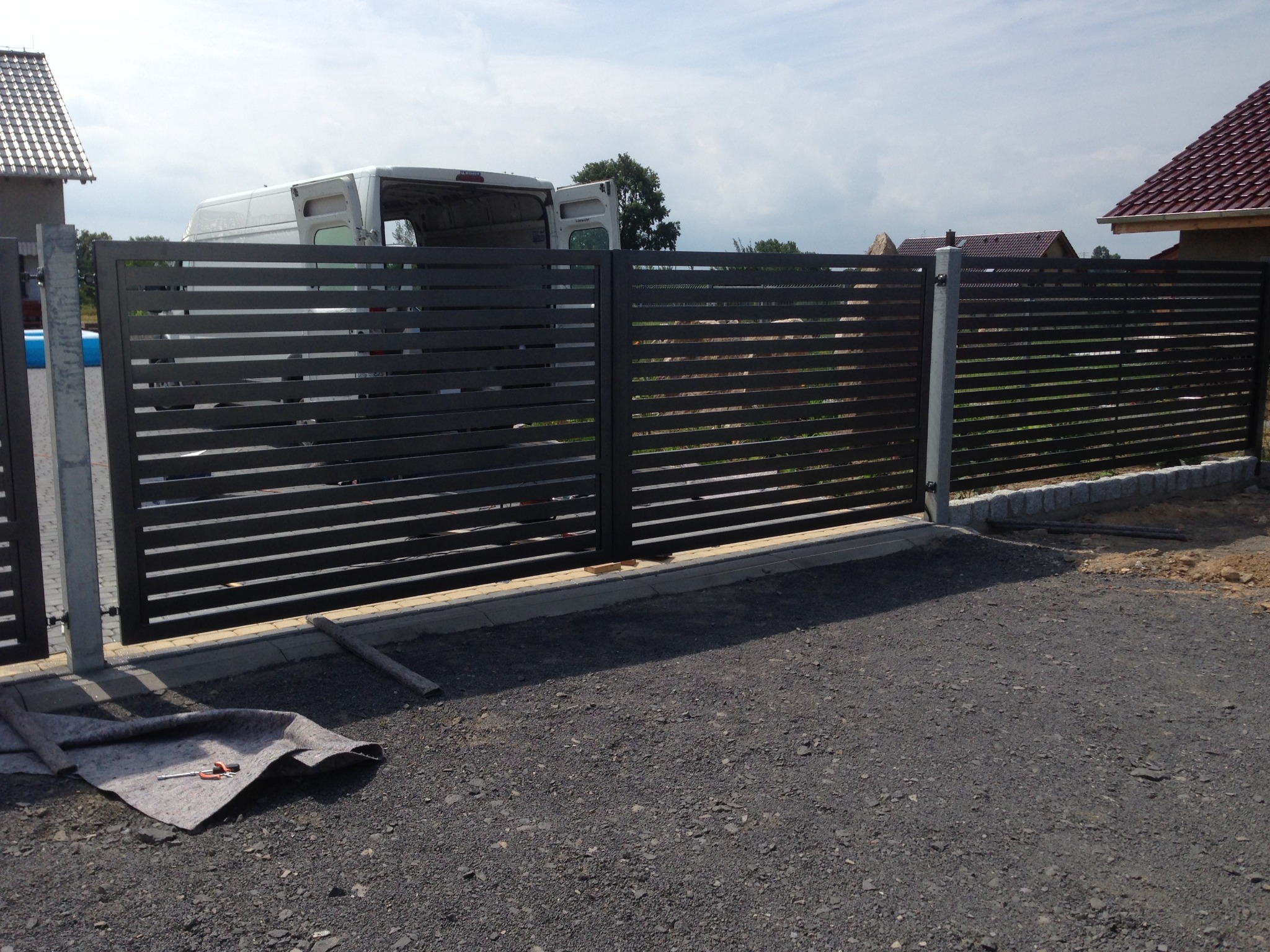 Nowoczesna, pozioma brama wjazdowa z metalowymi lamelami w kolorze antracytowym, widoczny częściowo biały bus z otwartymi drzwiami w tle, na podłożu mata ochronna i narzędzia.