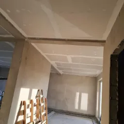 Wnętrze w trakcie prac wykończeniowych: zamontowane płyty na suficie i ścianach, widoczne łączenia płyt, drewniana paleta w rogu pomieszczenia, surowe ściany w tle, okno z widocznym światłem dziennym.