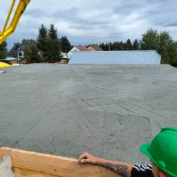 Wylewanie betonu na szalunek fundamentów za pomocą pompy, widoczna zbrojenia, w tle zabudowa jednorodzinna, pracownik w zielonym kasku z tatuażem na ramieniu.