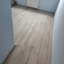 Jasne panele podłogowe ułożone w nowym pokoju z niebieskimi ścianami, widoczny grzejnik i parapet z kwiatami.