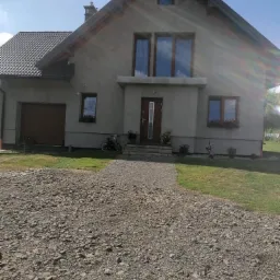 Surowy dom jednorodzinny z szarą elewacją w trakcie budowy, widoczne okna z drewnianymi ramami, szara dachówka i kamienista droga dojazdowa, dwa rowery dziecięce zaparkowane przy ścianie.