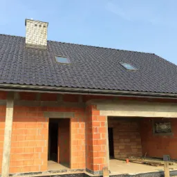 Dom w trakcie budowy, widoczne ściany z czerwonej cegły, zamontowany dach z ciemnej dachówki z oknami dachowymi i komin z jasnej cegły, rynny zamontowane.