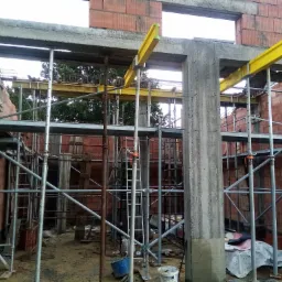 Wznoszenie ścian z czerwonej cegły na budowie domu, widoczne szalunki okienne z żółtymi belkami i metalowe rusztowania.