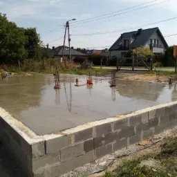 Zalany betonem fundament z wystającymi prętami zbrojeniowymi, otoczony murem z szarych bloczków betonowych, w tle domy jednorodzinne i słup energetyczny.