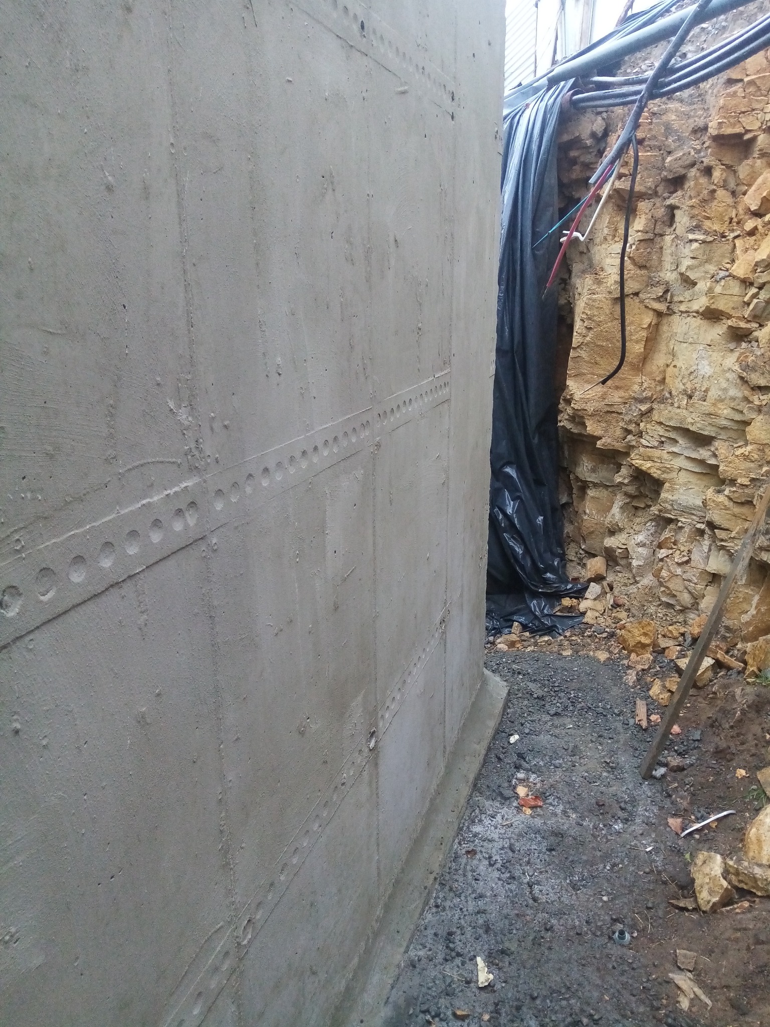 Betonowa ściana fundamentowa z poziomymi śladami po szalunku, widoczne rury i przewody elektryczne zabezpieczone folią oraz skaliste podłoże wykopu.