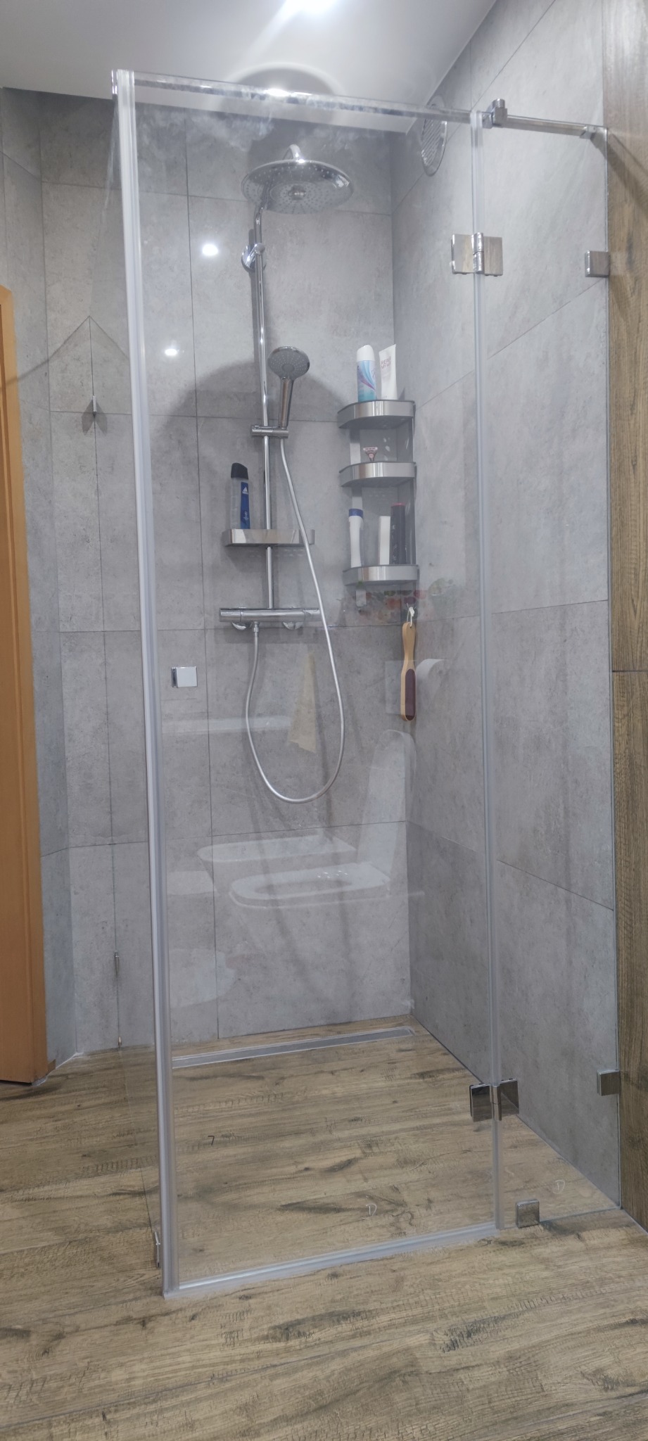 Kabina prysznicowa ze szklanymi drzwiami, widoczne chromowane zawiasy i odpływ liniowy w podłodze imitującej drewno, na ścianie szara płytka.