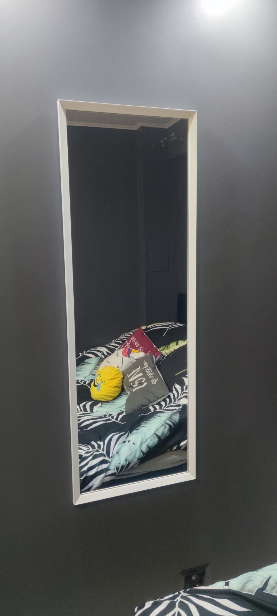 Prostokątne lustro w białej ramie zawieszone na szarej ścianie, odbijające fragment sypialni z wzorzystą pościelą i dekoracyjnymi poduszkami.