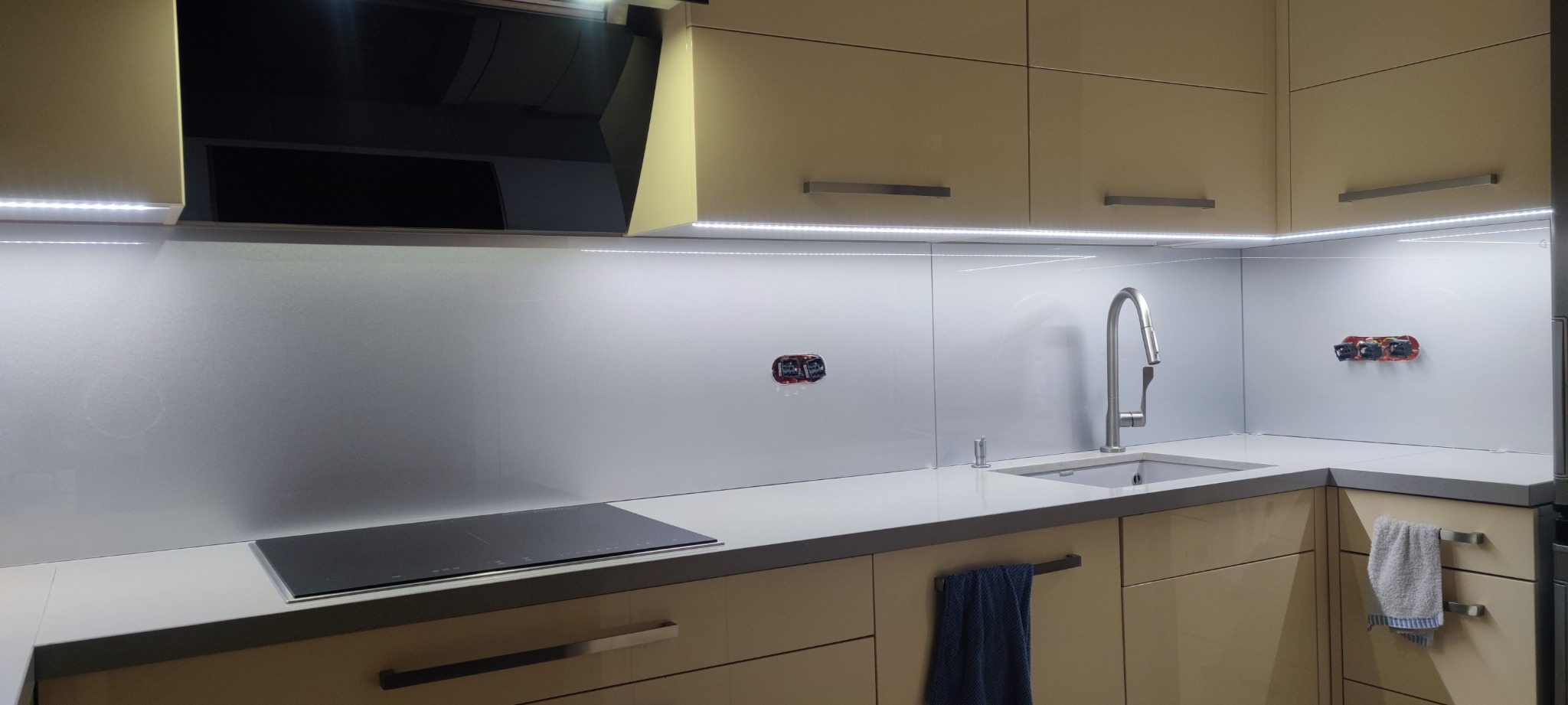 Kuchnia z jasnymi, lakierowanymi szafkami, szklanym panelem ściennym z oświetleniem LED i niedokończonymi gniazdkami elektrycznymi, nowoczesna bateria kuchenna.