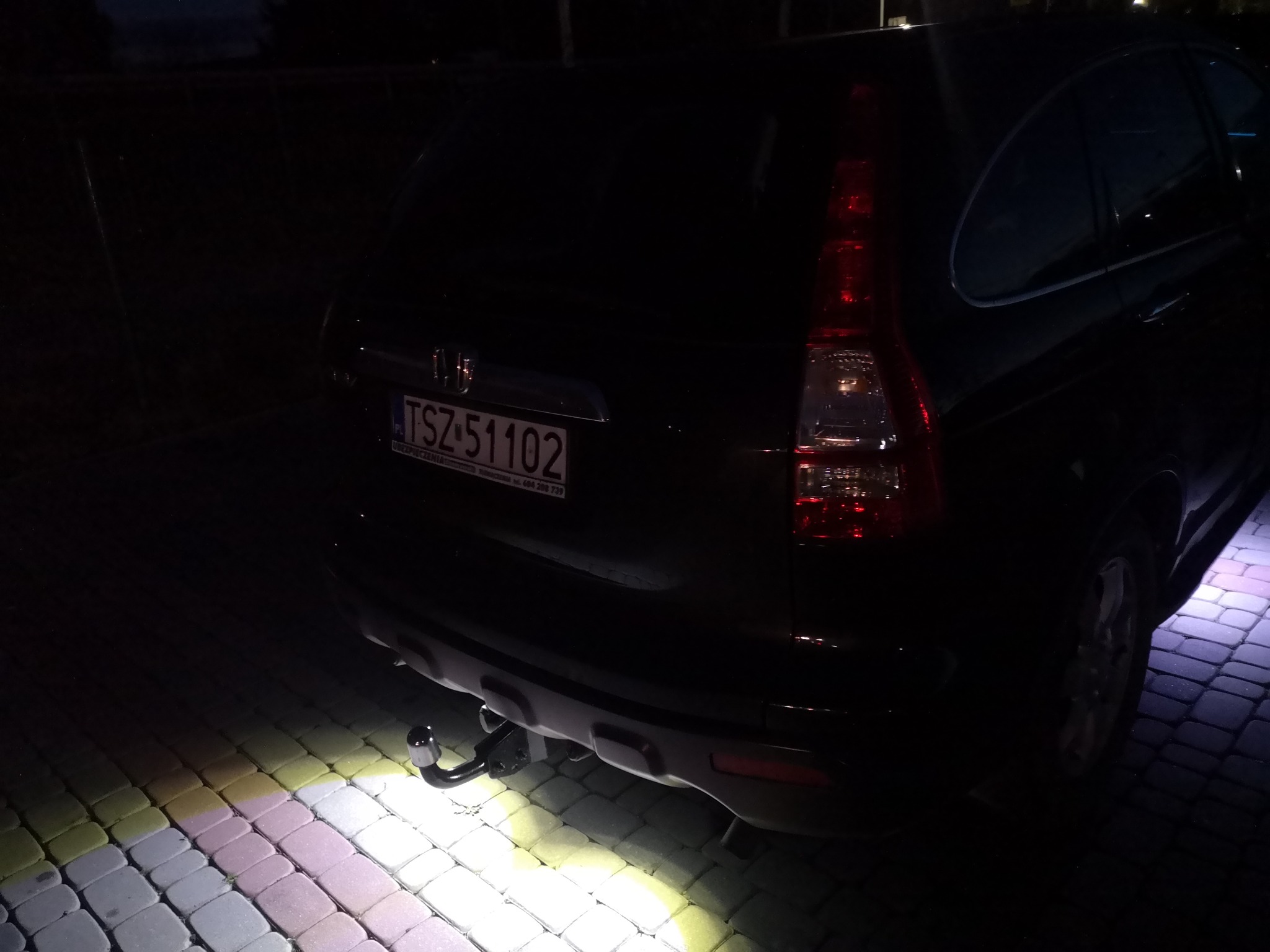 Częściowo oświetlony tył czarnego samochodu osobowego marki Honda z zamontowanym hakiem holowniczym, stojącego na kostce brukowej w słabym oświetleniu.