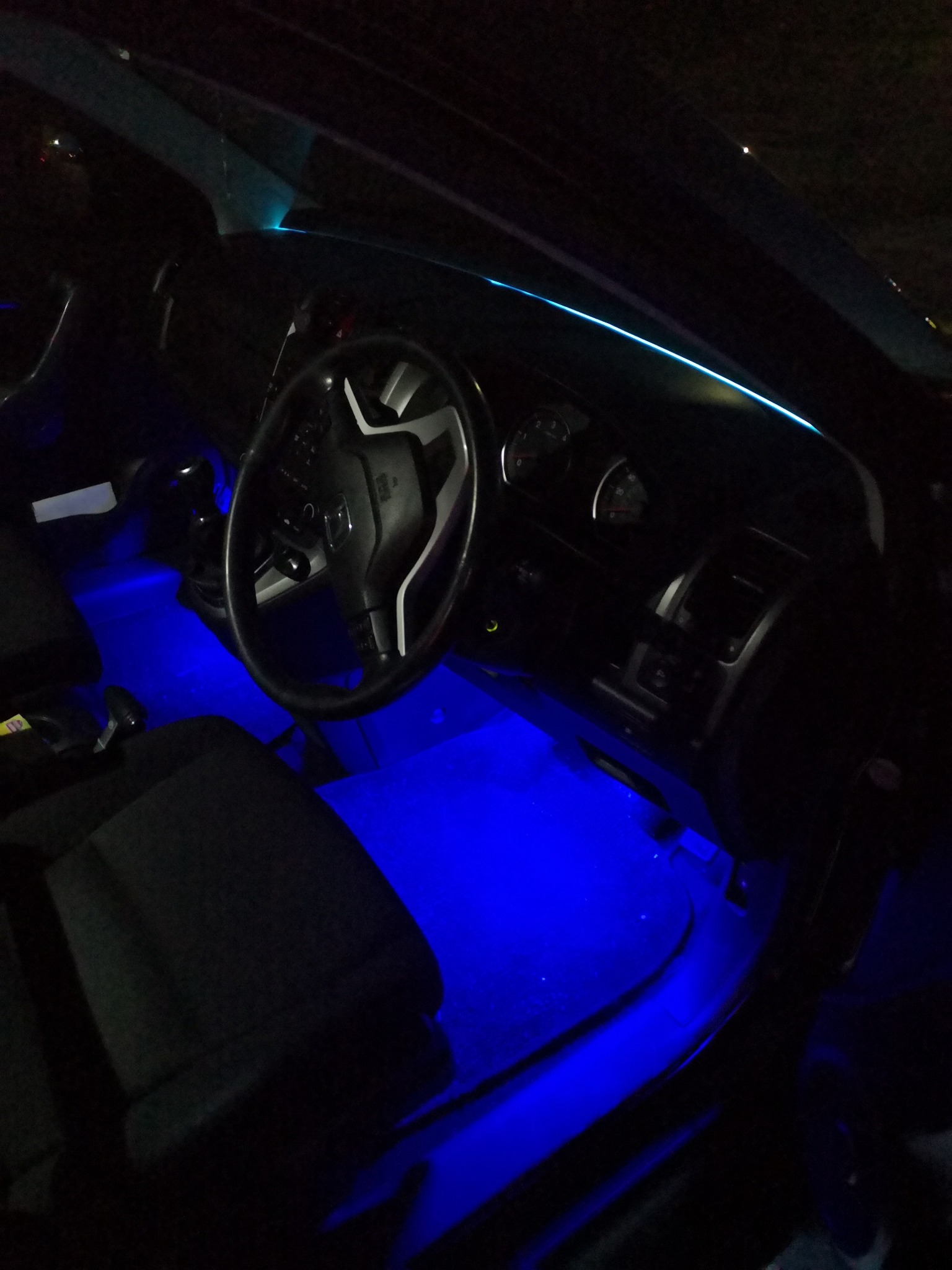 Wnętrze samochodu z podświetleniem LED w kolorze niebieskim na podłodze i desce rozdzielczej, widoczna kierownica i fragment deski rozdzielczej.