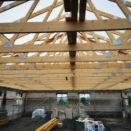 Wnętrze surowego domu z widoczną konstrukcją drewnianego dachu, ściany z betonu komórkowego, rusztowania i materiały budowlane na podłodze.