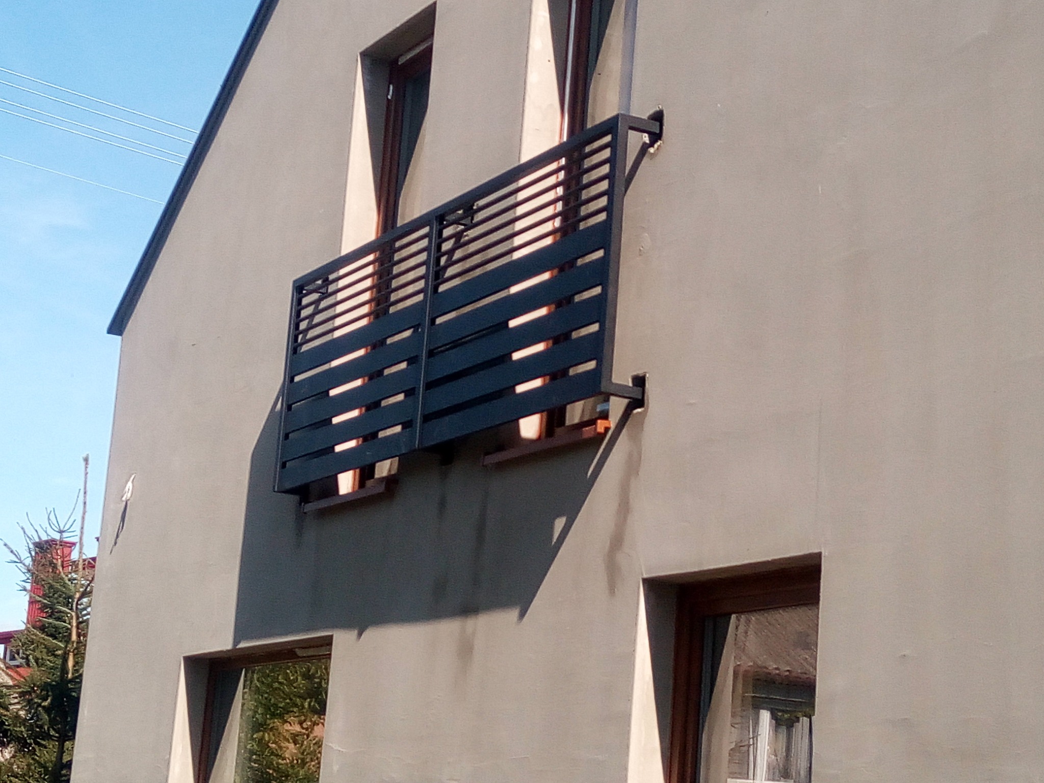 Czarna, pozioma balustrada balkonowa z metalowych profili, zamocowana do szarej ściany budynku z widocznymi oknami.