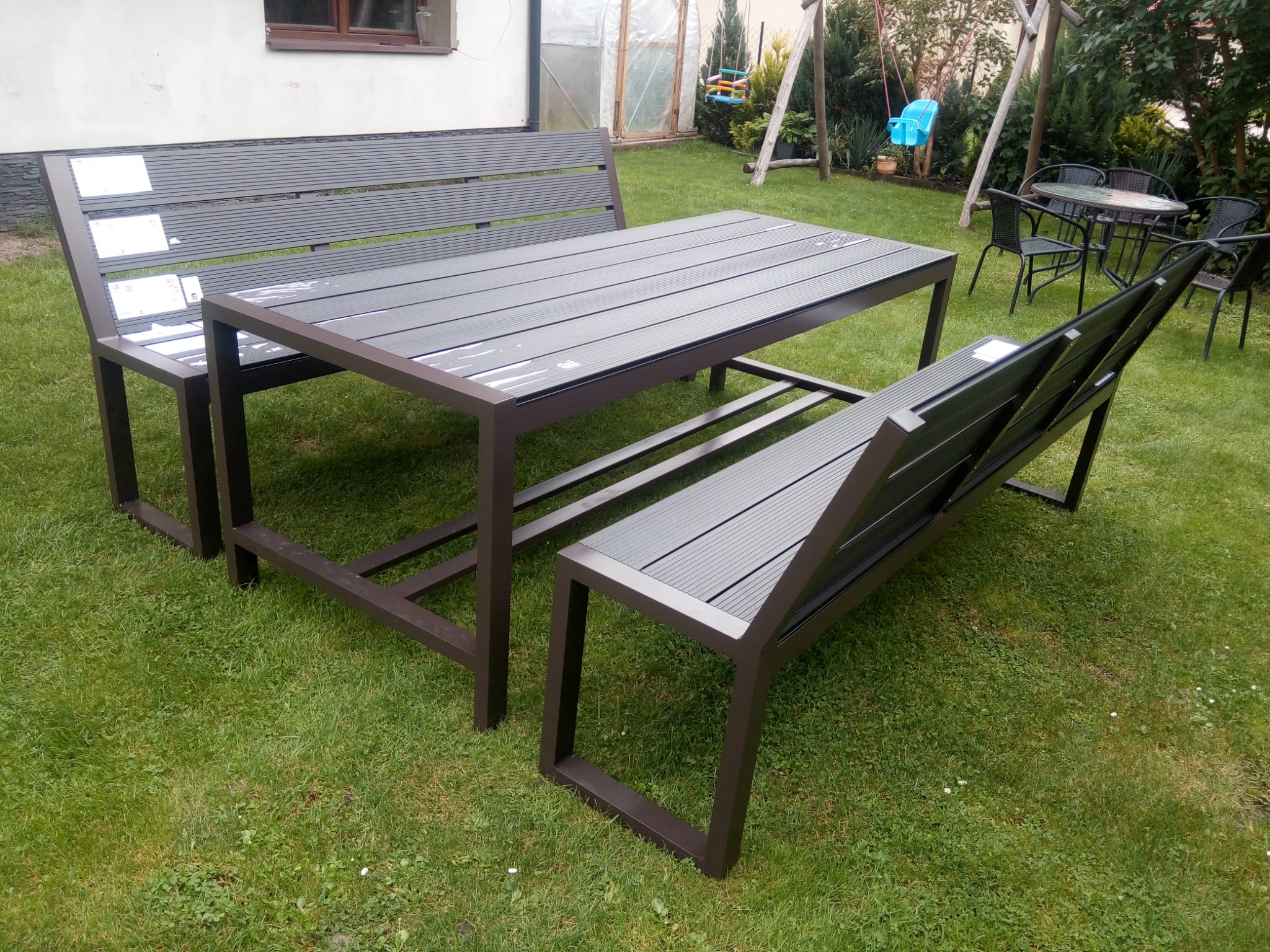 Zestaw mebli ogrodowych: stół i dwie ławki z metalową ramą w kolorze ciemnego brązu i szarymi siedziskami z tworzywa sztucznego, ustawiony na trawniku przed domem.