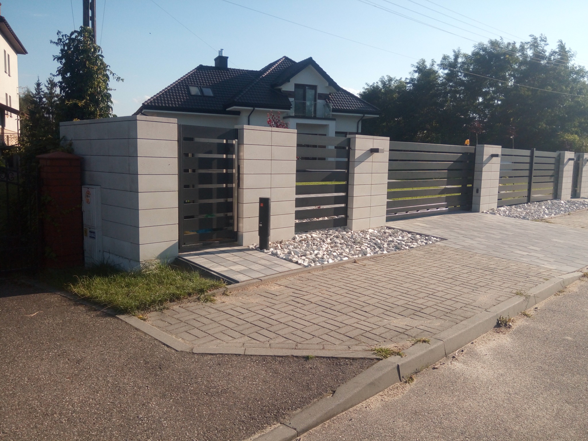 Nowoczesne ogrodzenie z poziomych paneli metalowych i betonowych słupków, uzupełnione białymi kamieniami dekoracyjnymi, prowadzące do domu z ciemnym dachem.
