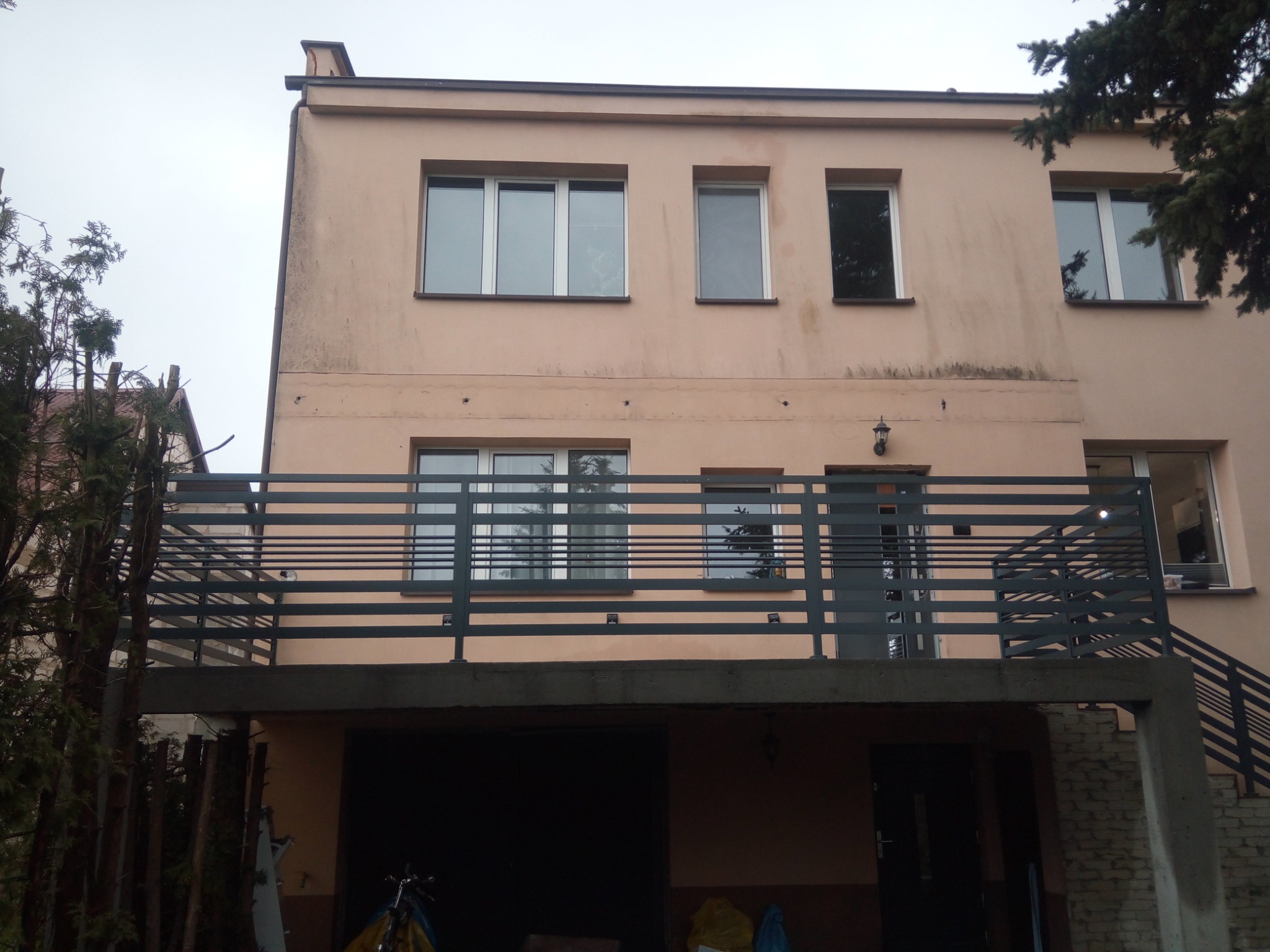 Metalowa balustrada balkonowa z poziomych profili na tle dwupiętrowego budynku z garażem, widok z dołu.