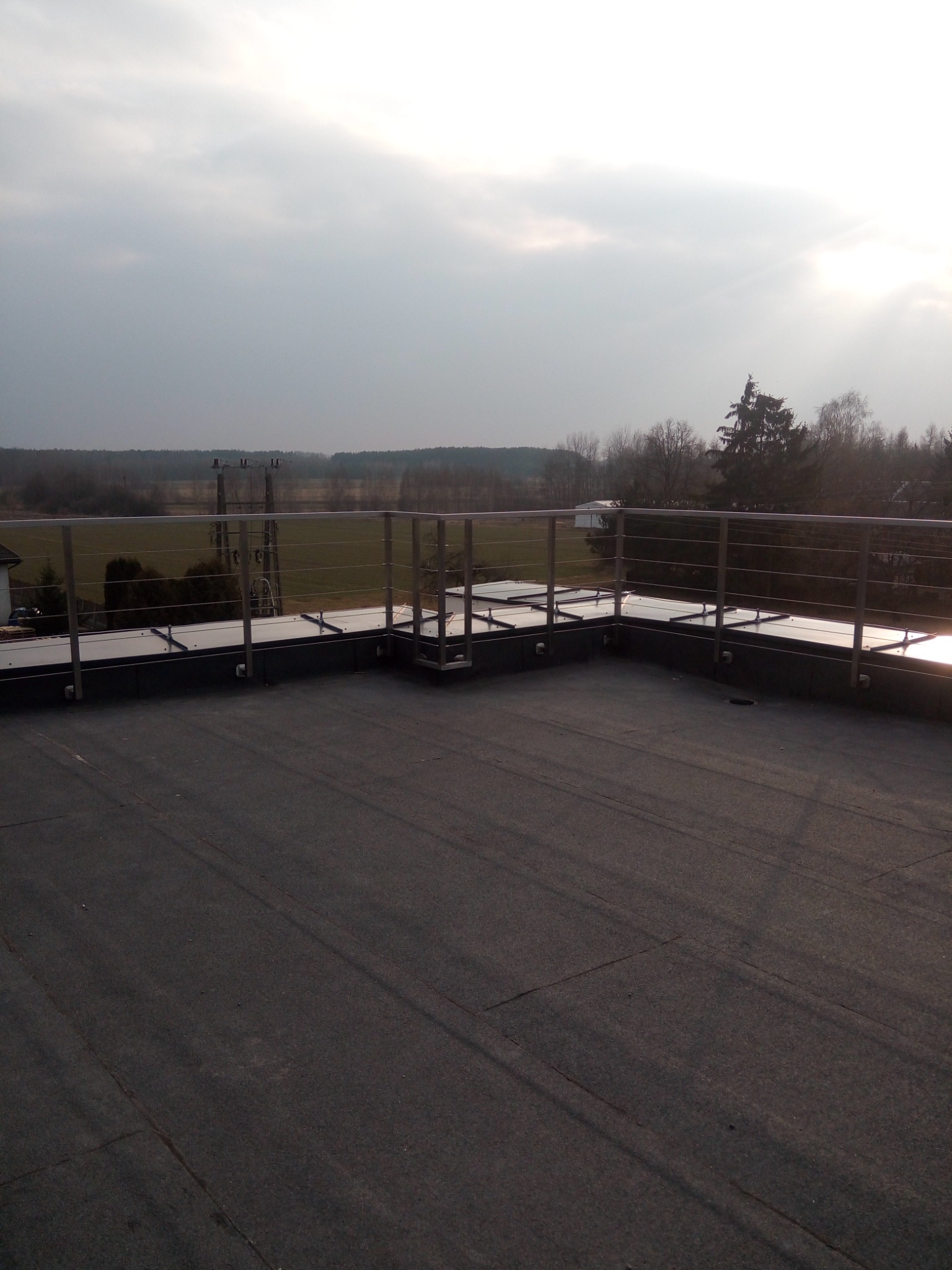 Metalowa balustrada z poziomymi linkami na dachu budynku, widok na zalesiony krajobraz w pochmurny dzień.