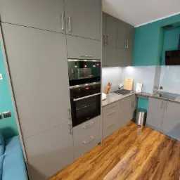 Szara zabudowa kuchenna z piekarnikiem i mikrofalówką, drewniana podłoga, turkusowa ściana w tle.