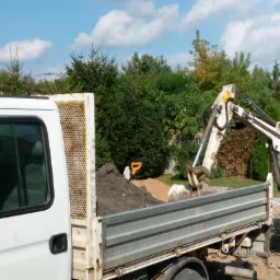 Mini koparka załadowuje urobek do skrzyni ładunkowej białego samochodu dostawczego na tle zieleni