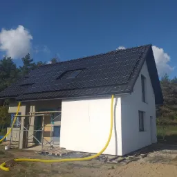 Dom jednorodzinny w trakcie ocieplania elewacji, widoczne rusztowania, żółte rury wentylacyjne i częściowo otynkowana fasada z panelami słonecznymi na dachu, w tle las i błękitne niebo z chmurami.