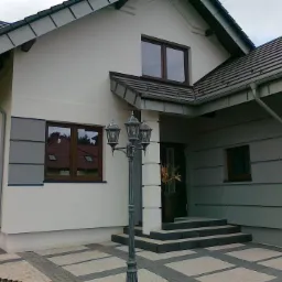 Dwukondygnacyjny dom jednorodzinny z biało-szarą elewacją, ciemną dachówką i stylizowaną latarnią przed wejściem, widok z poziomu chodnika wyłożonego kostką.