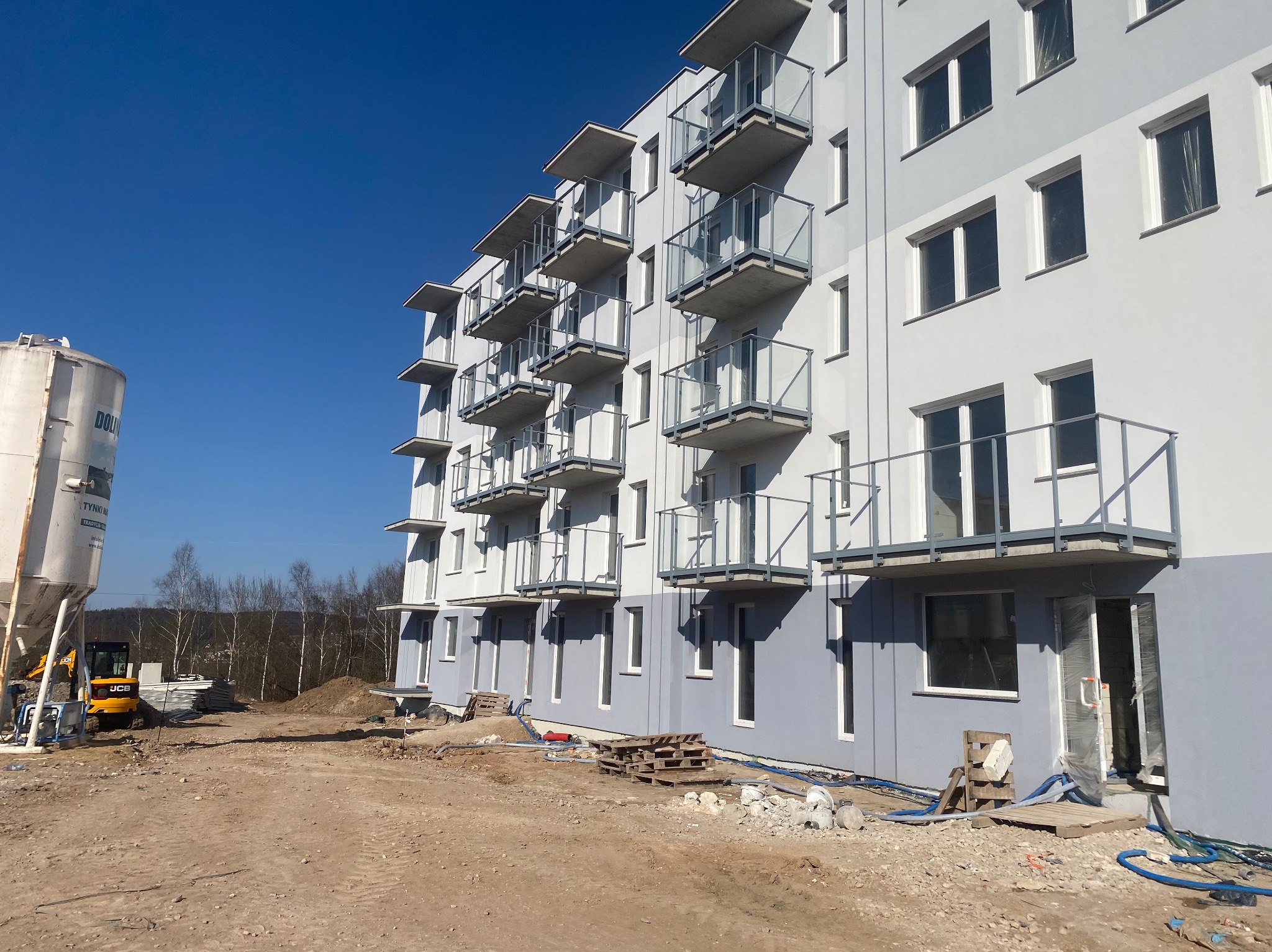 Nowo otynkowany, wielopiętrowy budynek mieszkalny z balkonami, widoczny z placu budowy z maszyną do tynkowania i stertą materiałów budowlanych.