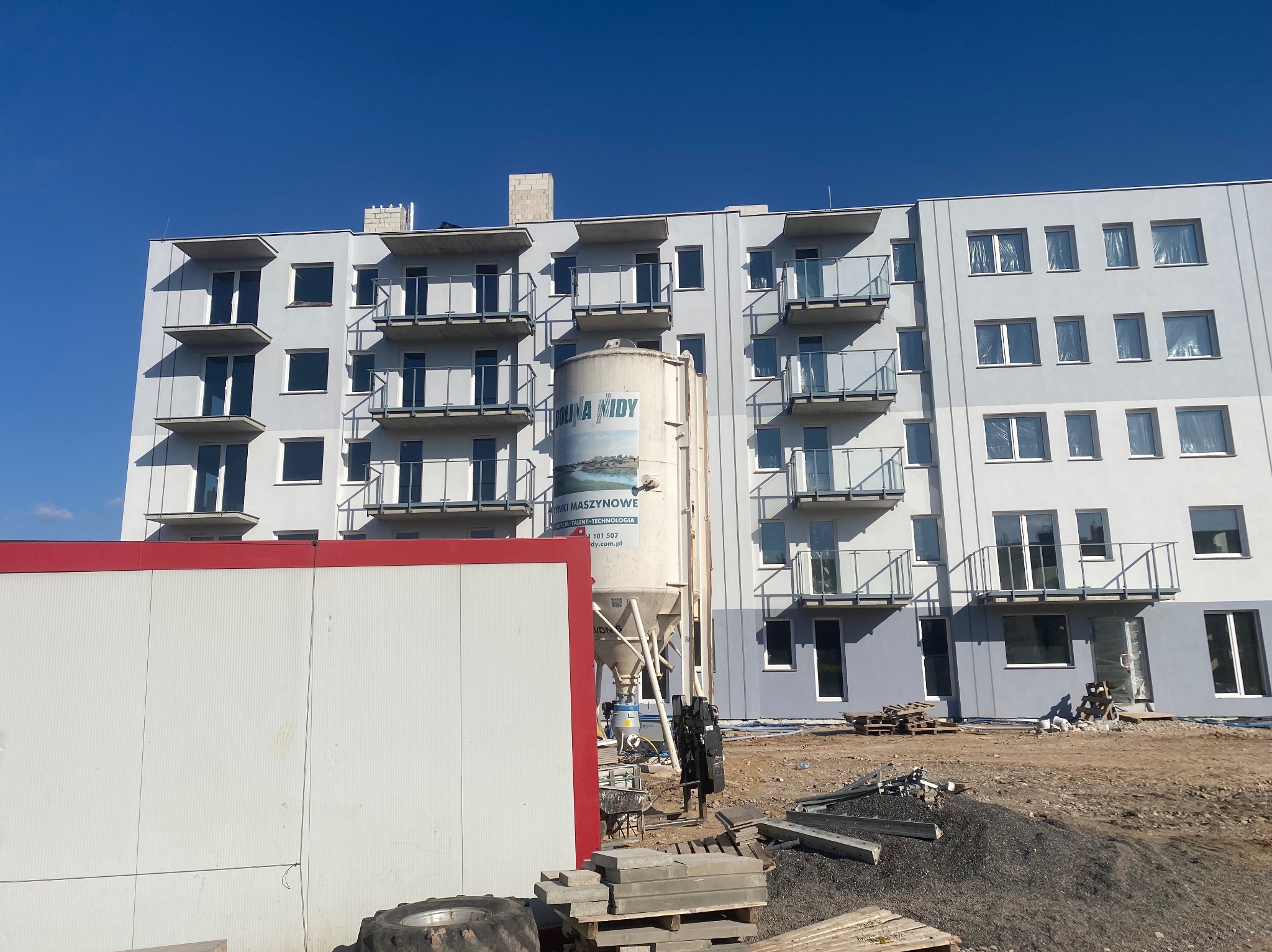 Nowo wybudowany budynek wielorodzinny z balkonami, widoczny zbiornik na zaprawę z logo firmy, teren budowy z materiałami i kontenerem biurowym.