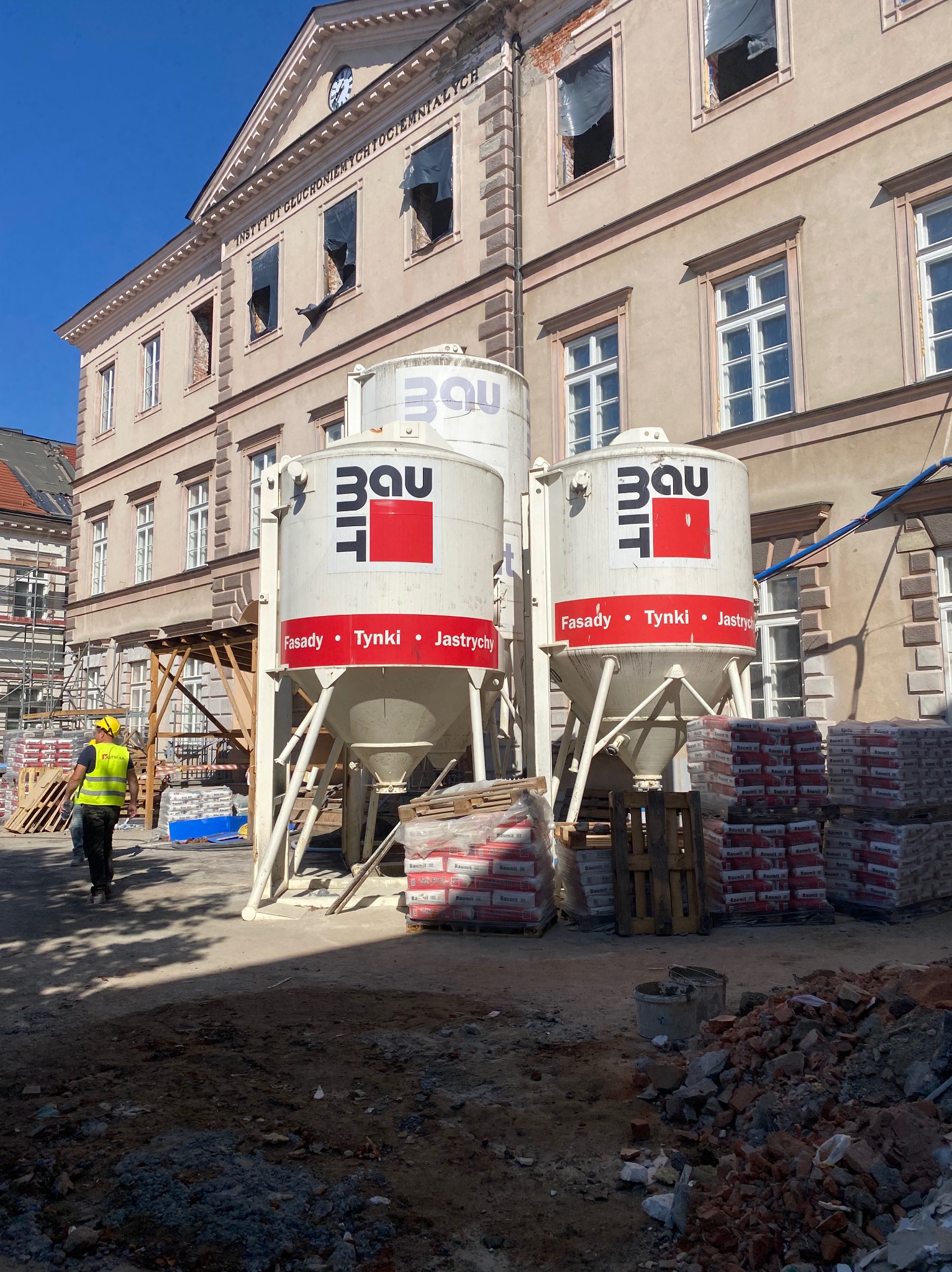Silosy z zaprawą Baumit na budowie w Kielcach, przygotowane do tynkowania maszynowego. Widoczny pracownik w kamizelce i worki z materiałami budowlanymi.