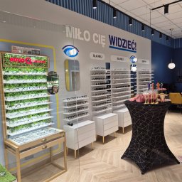 Piotr Krzeminski Pod Klucz - Wnętrze salonu optycznego z ekspozycją okularów, w tym regał z zielenią i okularami marki Ethnia Barcelona, oraz stolik z dekoracjami i napisem 'Miło Cię Widzieć!'.