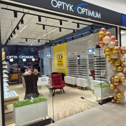 Piotr Krzeminski Pod Klucz - Fasada salonu optycznego Optyk Optimum z dekoracją balonową w odcieniach bieli, złota i różowego złota. Wnętrze widoczne przez szklane drzwi, z ekspozycją okularów, czerwonym fotelem i plakatem...