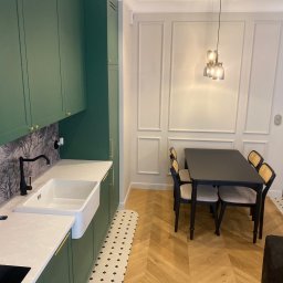 Piotr Krzeminski Pod Klucz - Nowoczesna kuchnia z zielonymi szafkami, białym zlewem farmerskim i blatem z wzorem. W tle stół z krzesłami i dekoracyjna ściana z panelami.